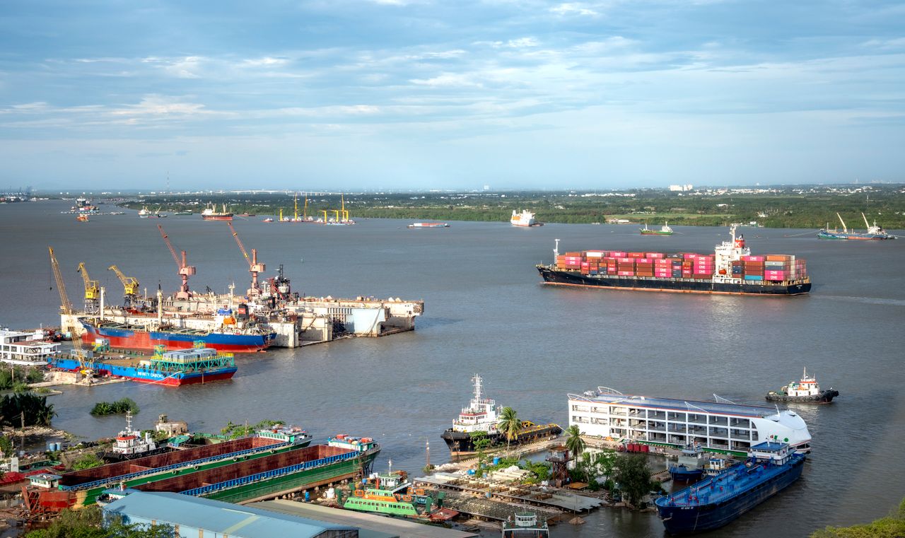 Home A busy shipping port with cargo ships and cranes under a clear sky, viewed from above.