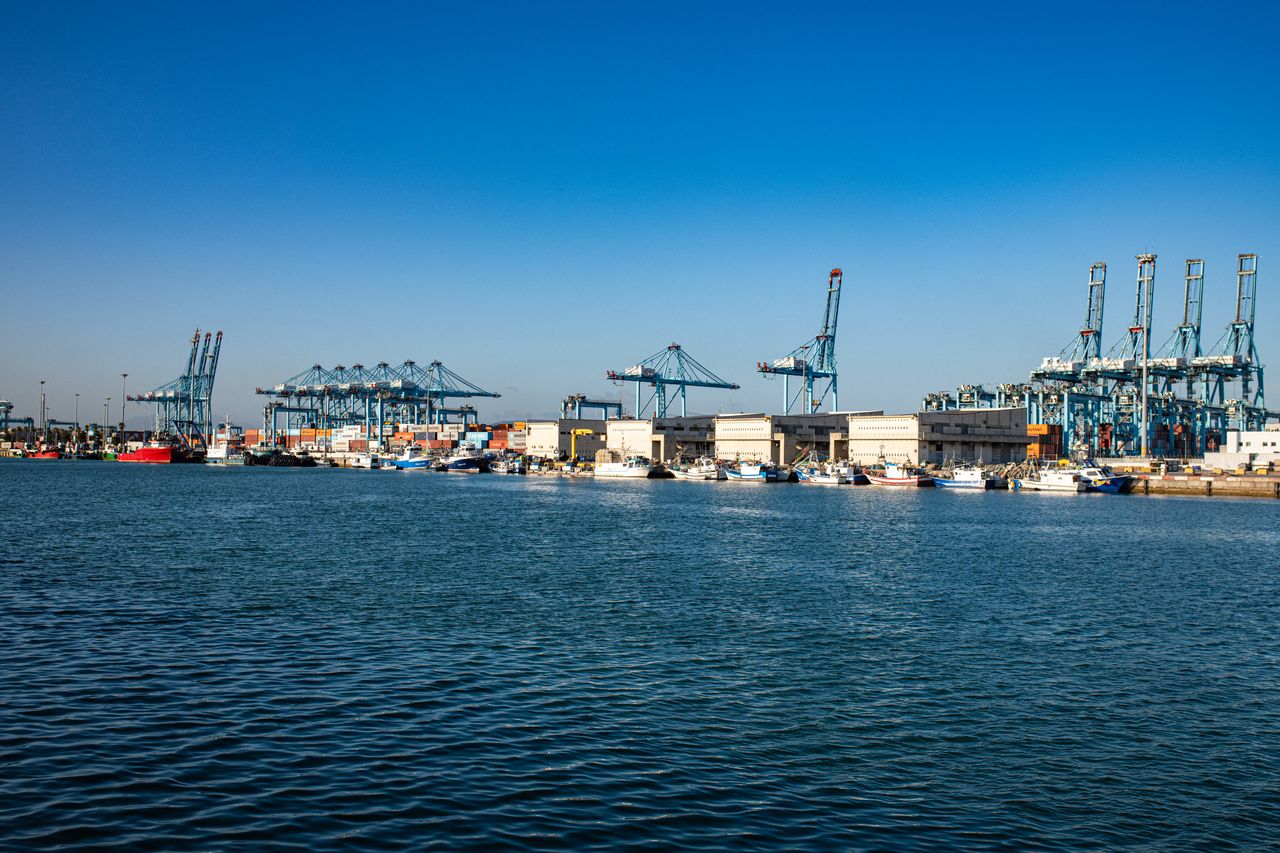 A bustling harbor with large cranes, cargo containers, and ships under a clear blue sky.